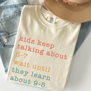 May include: Cream-coloured t-shirt with the text "kids keep talking about 6-7 wait until they learn about 9-5" in pink, yellow, and blue. The shirt is paired with light-wash jeans, a tan hat, and a necklace.