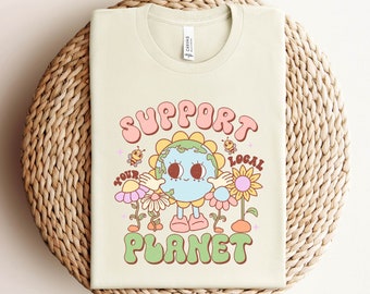 May include: A light beige t-shirt with a graphic of a cartoon Earth holding a flower. The text "Support Your Planet" is written in a retro font above the Earth and "Local" is written in a retro font below the Earth.
