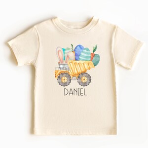 Könnte beinhalten: Ein cremefarbenes T-Shirt mit einem gelben Kipplaster, der Ostereier und einen Hasen transportiert. Der Text "DANIEL" ist unter dem Laster gedruckt.