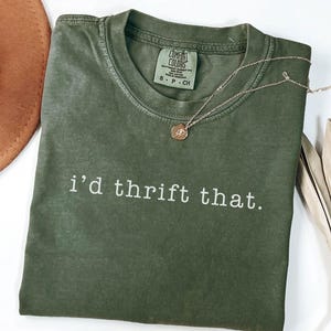 Pode incluir: Uma camiseta verde-oliva com a frase "i'd thrift that" impressa em branco. Um colar de ouro com um pingente repousa sobre a camiseta. Um chapéu marrom e uma planta seca estão no fundo.