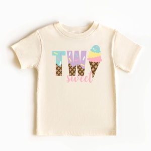 May include: A light beige t-shirt with the words "TWO sweet" printed on it. The "TWO" is made up of ice cream cones with sprinkles and the word "sweet" is in pink.