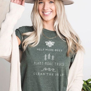 K&ouml;nnte beinhalten: Ein gr&uuml;nes T-Shirt mit dem Text "Help More Bees, Plant More Trees, Clean The Seas" und einer Bienengrafik. Das Shirt wird von einer Person mit blonden Haaren und einem braunen Hut getragen.