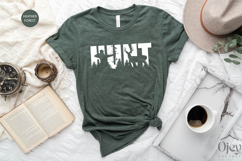 May include: A green t-shirt with the word "HUNT" in white text. The letters are formed by silhouettes of a hunter with a bow and arrow, trees, and a deer.