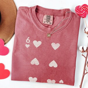 May include: A dusty rose t-shirt with a six of hearts design. The shirt features a crew neck and a small tag at the neckline. A silver necklace with a pendant rests on the shirt. A heart-shaped lollipop and cotton branch are also in the image.