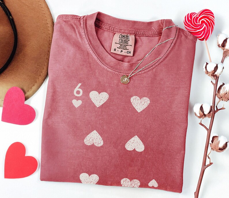 May include: A dusty rose t-shirt with a six of hearts design. The shirt features a crew neck and a small tag at the neckline. A silver necklace with a pendant rests on the shirt. A heart-shaped lollipop and cotton branch are also in the image.