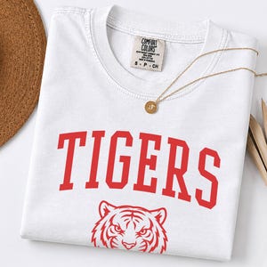 May include: A white short-sleeved t-shirt featuring the word TIGERS in large red collegiate-style lettering above a red graphic illustration of a tiger face. The shirt has a Comfort Colors brand label at the inner neckline and a crew neck collar.