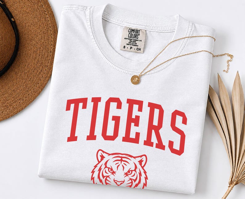 May include: A white short-sleeved t-shirt featuring the word TIGERS in large red collegiate-style lettering above a red graphic illustration of a tiger face. The shirt has a Comfort Colors brand label at the inner neckline and a crew neck collar.