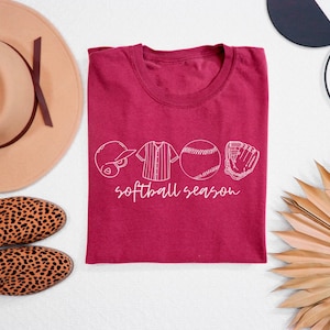 May include: A maroon t-shirt with a white graphic design of a baseball, glove, bat, and jersey with the text "softball season".