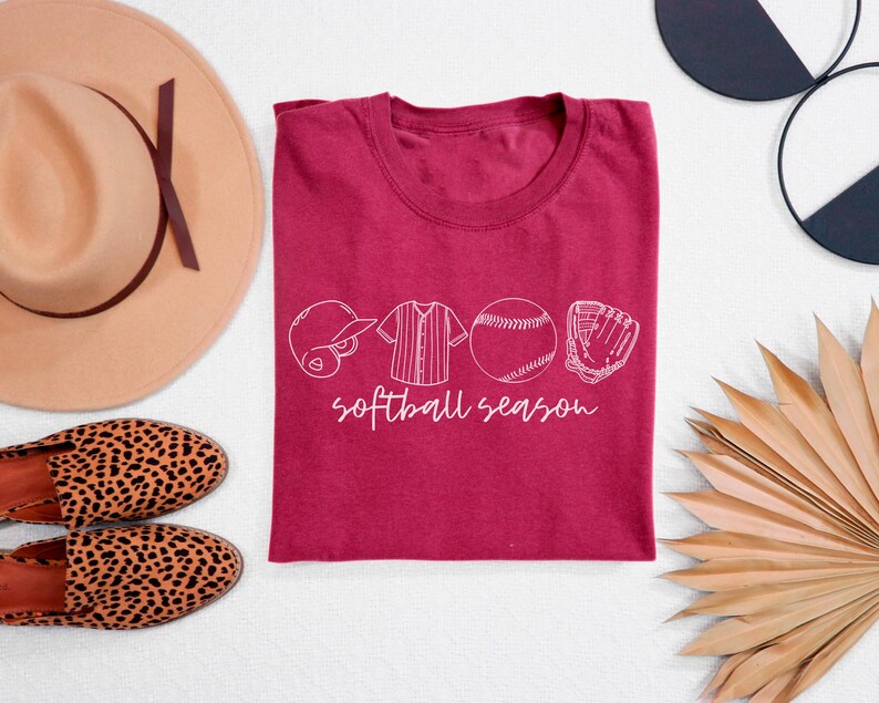 May include: A maroon t-shirt with a white graphic design of a baseball, glove, bat, and jersey with the text "softball season".