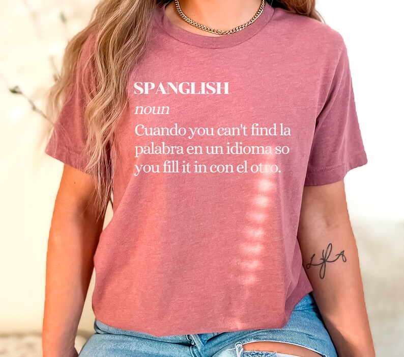Puede incluir: Una camiseta color malva con la palabra "SPANGLISH" definida como "Cuando you can't find la palabra en un idioma so you fill it in con el otro."