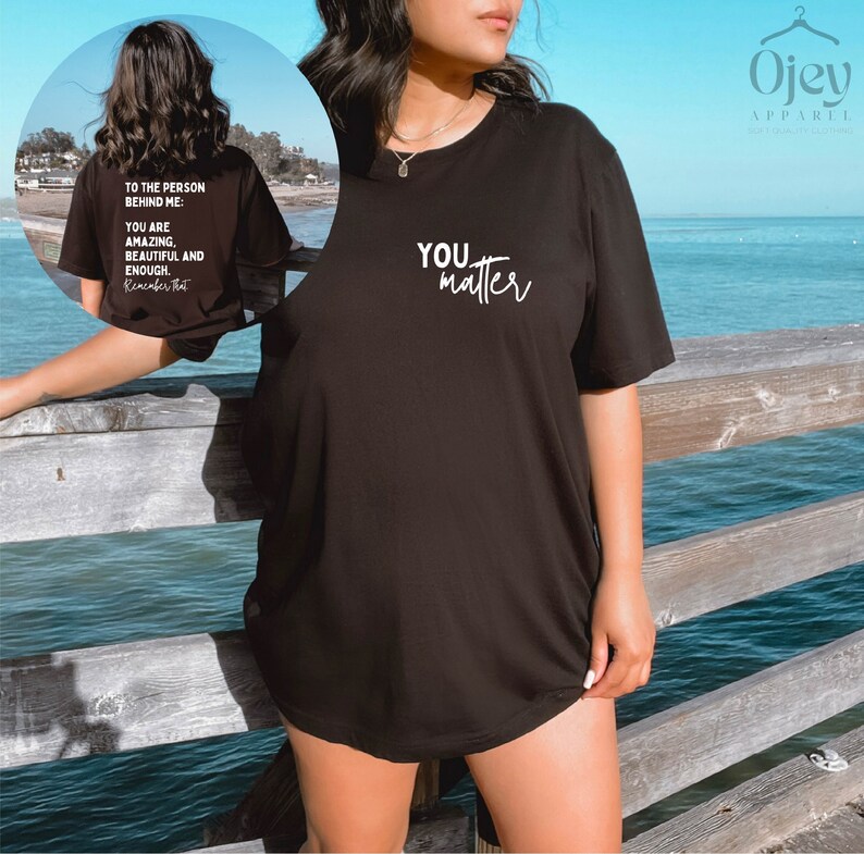 May include: Black oversized t-shirt with the text "YOU matter" in white cursive lettering. The shirt is worn by a person standing on a wooden pier overlooking the ocean.