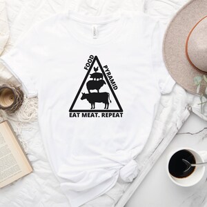 May include: White t-shirt with a black graphic of a food pyramid with a cow, sheep, and pig on top. The text "Food Pyramid" is on the right side of the triangle and the text "Eat Meat. Repeat" is at the bottom.