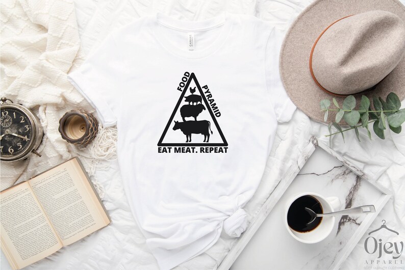 May include: White t-shirt with a black graphic of a food pyramid with a cow, sheep, and pig on top. The text "Food Pyramid" is on the right side of the triangle and the text "Eat Meat. Repeat" is at the bottom.