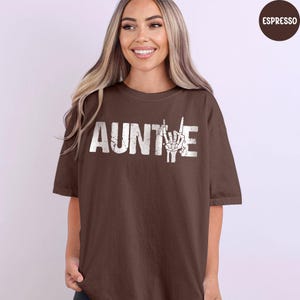 May include: A brown t-shirt with the word "AUNTIE" in white block letters and a white hand gesture graphic. The shirt is a solid color and has short sleeves.