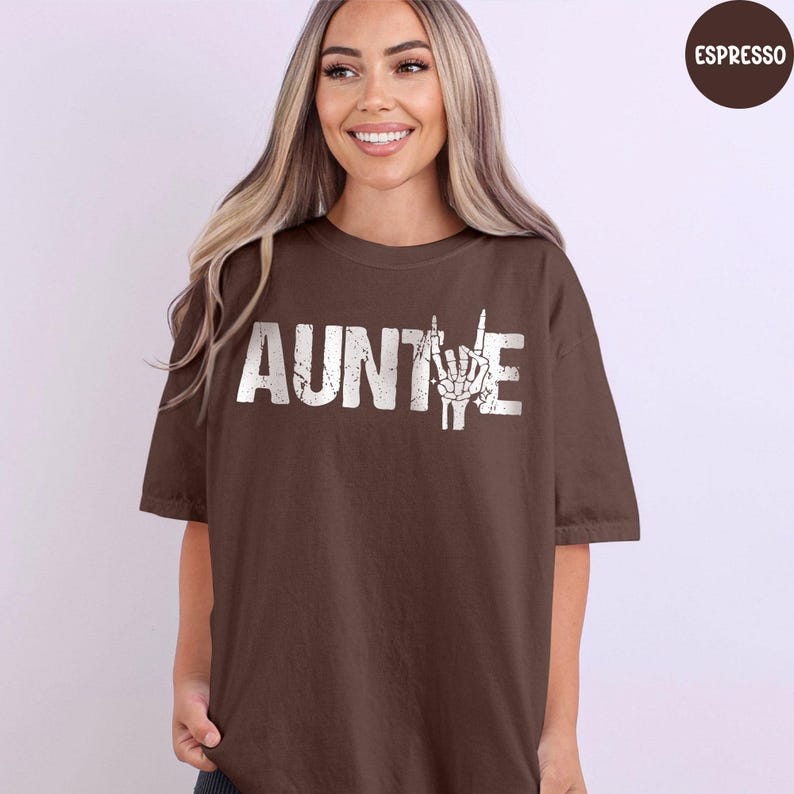 May include: A brown t-shirt with the word "AUNTIE" in white block letters and a white hand gesture graphic. The shirt is a solid color and has short sleeves.