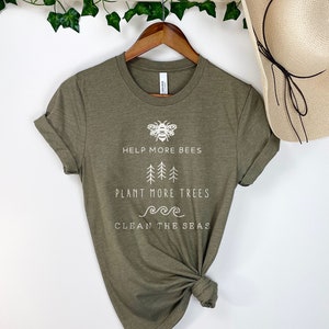 K&ouml;nnte beinhalten: Ein gr&uuml;nes T-Shirt mit wei&szlig;em Text, der "Help More Bees, Plant More Trees, Clean The Seas" lautet. Der Text ist von einfachen Illustrationen einer Biene, B&auml;umen und Wellen umgeben.