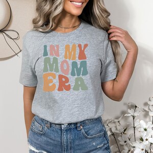 May include: A gray t-shirt with the text "In My Mom Era" in colorful, retro-style lettering.