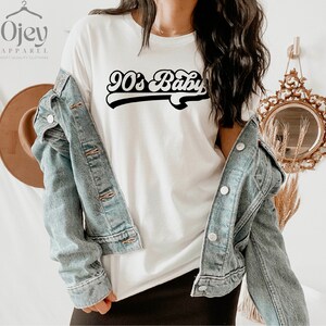 Op de afbeelding: Wit T-shirt met een zwarte retro-afbeelding met de tekst "90's Baby". Het model draagt een spijkerjack en een zwarte rok.