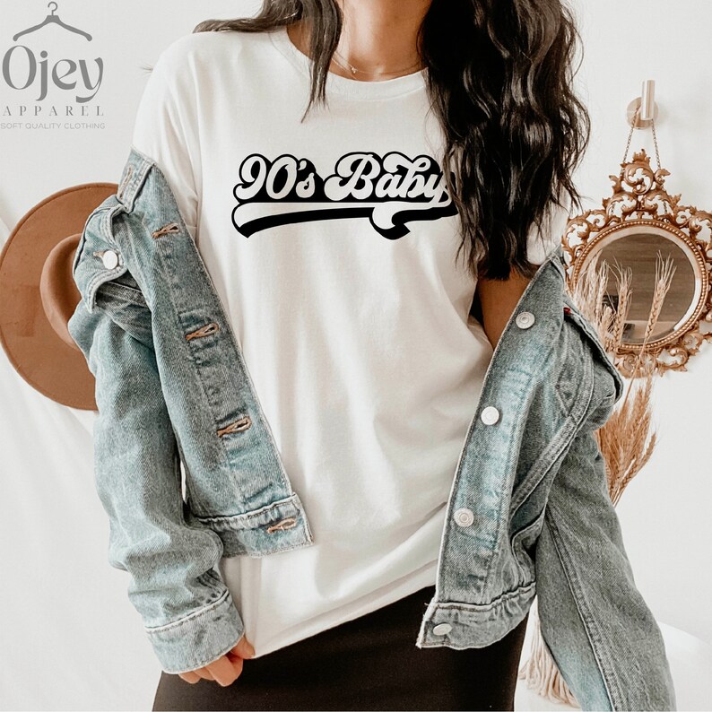 Op de afbeelding: Wit T-shirt met een zwarte retro-afbeelding met de tekst "90's Baby". Het model draagt een spijkerjack en een zwarte rok.