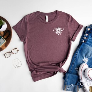 May include: A maroon t-shirt with a white bee graphic on the front. The shirt is laid out on a white surface with a pair of jeans, a pair of white sneakers, a plant, a camera, and a pair of glasses.
