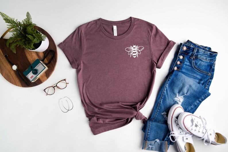 May include: A maroon t-shirt with a white bee graphic on the front. The shirt is laid out on a white surface with a pair of jeans, a pair of white sneakers, a plant, a camera, and a pair of glasses.