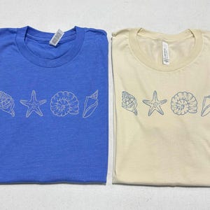 May include: Two t-shirts, one blue and one beige, each with a line of white seashell graphics. The designs include a conch shell, starfish, spiral shell, and another conch shell. The shirts are folded and ready to wear.