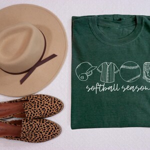May include: A green t-shirt with a white graphic of a baseball, a baseball glove, a baseball cap, and a baseball jersey. The text "softball season" is printed below the graphic.