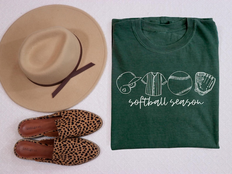 May include: A green t-shirt with a white graphic of a baseball, a baseball glove, a baseball cap, and a baseball jersey. The text "softball season" is printed below the graphic.