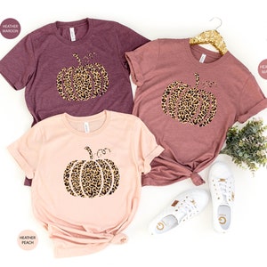 May include: Three t-shirts with a leopard print pumpkin design. The shirts are in heather maroon, heather mauve, and heather peach.