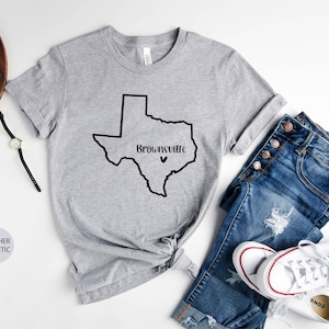 Op de afbeelding: Grijs T-shirt met een zwarte omtrek van de staat Texas en de tekst "Brownsville" met een klein hartje erboven.