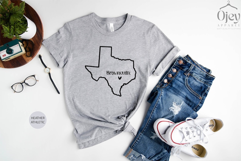 Op de afbeelding: Grijs T-shirt met een zwarte omtrek van de staat Texas en de tekst "Brownsville" met een klein hartje erboven.
