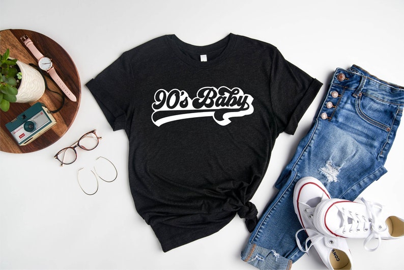 Op de afbeelding: Een zwart T-shirt met witte tekst die "90's Baby" zegt in een retro-lettertype. Het shirt ligt op een wit oppervlak met een spijkerbroek, een paar witte sneakers, een horloge, een camera en een bril.