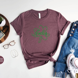 May include: Heather maroon t-shirt with a green shamrock and the word "lucky" written inside. The shirt is laid flat on a white surface with a pair of jeans, white sneakers, and other accessories.
