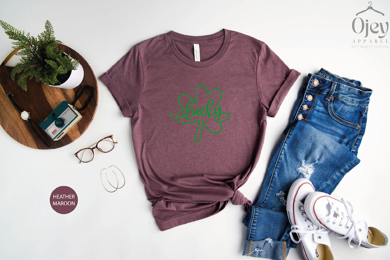 May include: Heather maroon t-shirt with a green shamrock and the word "lucky" written inside. The shirt is laid flat on a white surface with a pair of jeans, white sneakers, and other accessories.