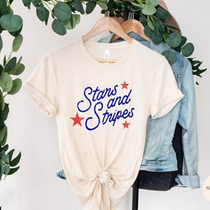 May include: A beige t-shirt with the text "Stars and Stripes" in blue with red stars printed on the front.