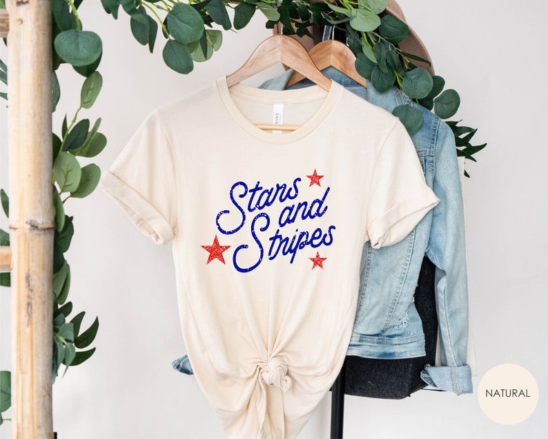 May include: A beige t-shirt with the text "Stars and Stripes" in blue with red stars printed on the front.