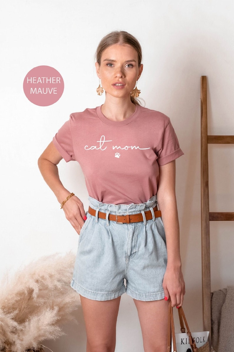 May include: A woman wearing a heather mauve t-shirt with the text "cat mom" and a white paw print. She is wearing denim shorts and a brown belt.