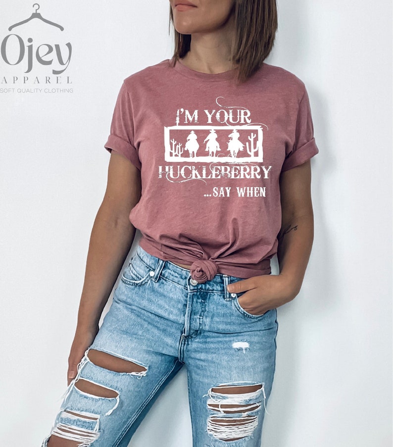 K&ouml;nnte beinhalten: Eine Frau tr&auml;gt ein staubiges rosafarbenes T-Shirt mit dem Text "I'm your huckleberry... say when" in Wei&szlig;. Das Shirt zeigt eine Grafik mit drei Cowboys, die auf Pferden reiten. Sie tr&auml;gt eine blaue Jeans mit Destroyed-Details.