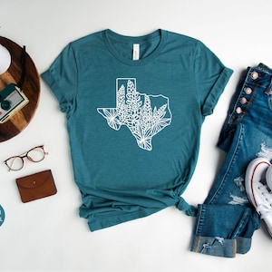May include: A teal t-shirt featuring a white outline of the state of Texas filled with floral designs. The shirt is styled with blue jeans and white sneakers. The words "OjeY Apparel" are in the top right corner.
