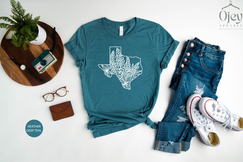 May include: A teal t-shirt featuring a white outline of the state of Texas filled with floral designs. The shirt is styled with blue jeans and white sneakers. The words "OjeY Apparel" are in the top right corner.