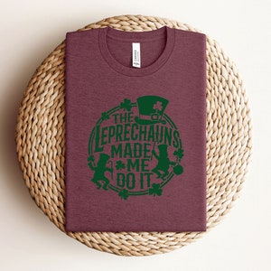 May include: A maroon t-shirt with a green graphic design that says "The Leprechauns Made Me Do It". The graphic features two leprechauns with green hats and green shamrocks.