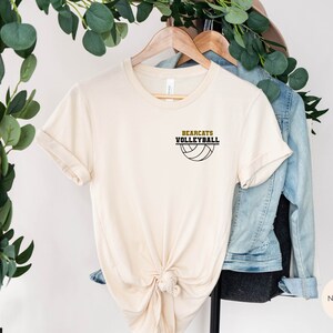 May include: A cream-coloured t-shirt featuring the text "BEARCATS VOLLEYBALL" above a volleyball graphic. The shirt is displayed on a wooden hanger, with a denim jacket and foliage in the background.