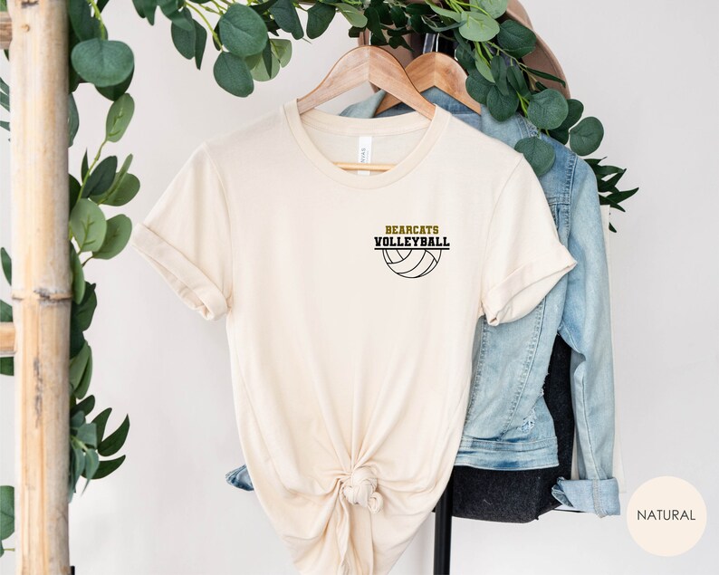 May include: A cream-coloured t-shirt featuring the text "BEARCATS VOLLEYBALL" above a volleyball graphic. The shirt is displayed on a wooden hanger, with a denim jacket and foliage in the background.