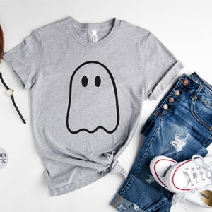 May include: A heather grey t-shirt with a black ghost graphic. The shirt is shown on a white background with a pair of jeans, a watch, and a pair of glasses.