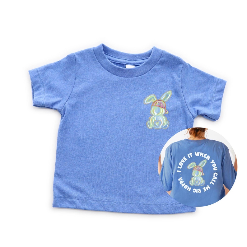 Puede incluir: Una camiseta de manga corta azul claro con un conejito de dibujos animados con una gorra de b&eacute;isbol al rev&eacute;s en la parte delantera. La parte posterior de la camiseta tiene un c&iacute;rculo blanco con el texto "I LOVE IT WHEN YOU CALL ME BIG HOPPA" y un conejito de dibujos animados con una gorra de b&eacute;isbol al rev&eacute;s.