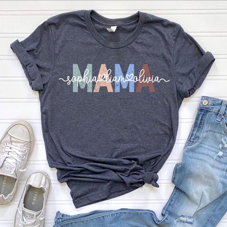 May include: Dark gray t-shirt with the word "MAMA" in colorful block letters. Below the word "MAMA" are the names "Sophia, Liam, Olivia" in a cursive font. The shirt is paired with blue jeans and gray sneakers.