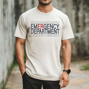 Może przedstawiać: Beżowa koszulka z nadrukiem "EMERGENCY DEPARTMENT" z przodu w kolorze białym i niebieskim, z wyr&oacute;żnioną czerwoną literą "E" w słowie "EMERGENCY".
