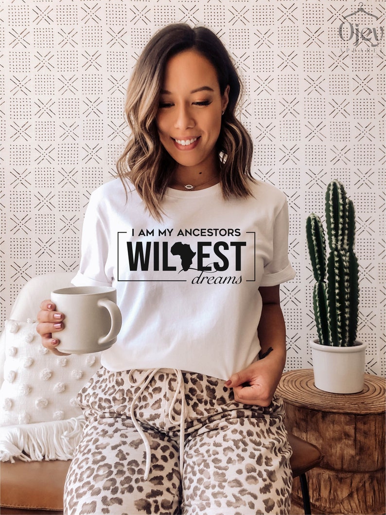 Peut inclure: T-shirt blanc avec un motif noir qui dit "I am my ancestors wildest dreams" avec un contour du continent africain. La personne portant le t-shirt tient une tasse blanche.