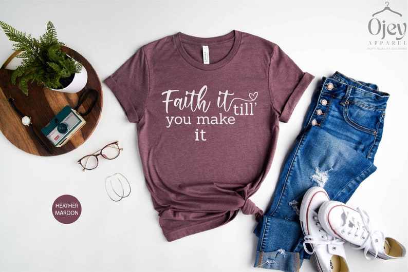 K&ouml;nnte beinhalten: Ein T-Shirt in Heide-Maroon mit dem Text "Faith it till' you make it" in wei&szlig;en Buchstaben. Das Shirt liegt flach auf einer wei&szlig;en Oberfl&auml;che mit einer Jeans, einem Paar wei&szlig;en Sneakers und einem Holztablett mit einer Kamera und einer Brille.