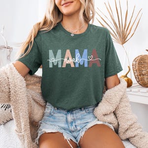 May include: A dark green t-shirt with the word "MAMA" in pastel colors and the name "Noah" in a cursive font. A small heart is next to the name. The shirt is worn with denim shorts and a cream-colored cardigan.